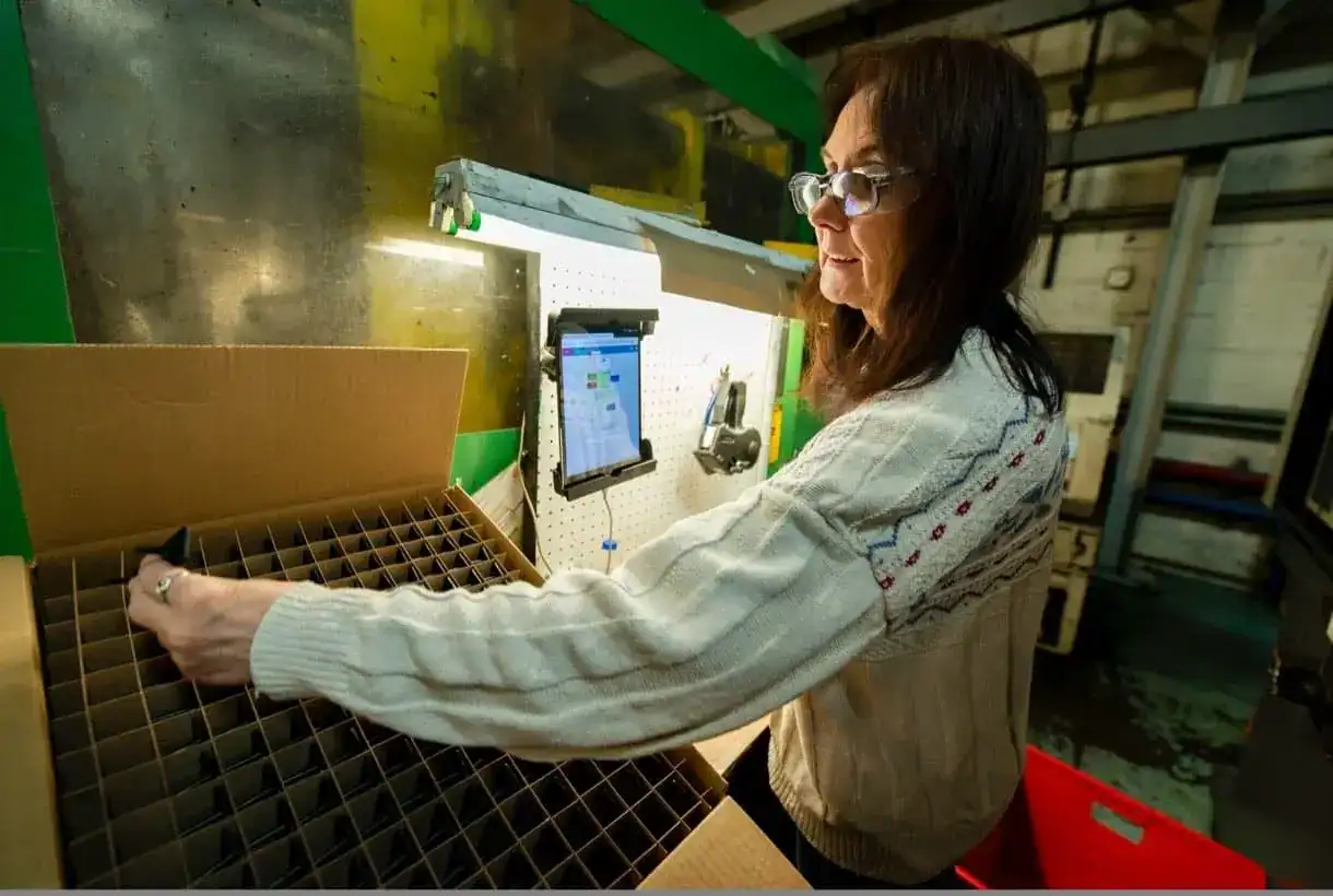 Woman packing parts into a divided carton at a station with a mounted tablet in a plastics factory