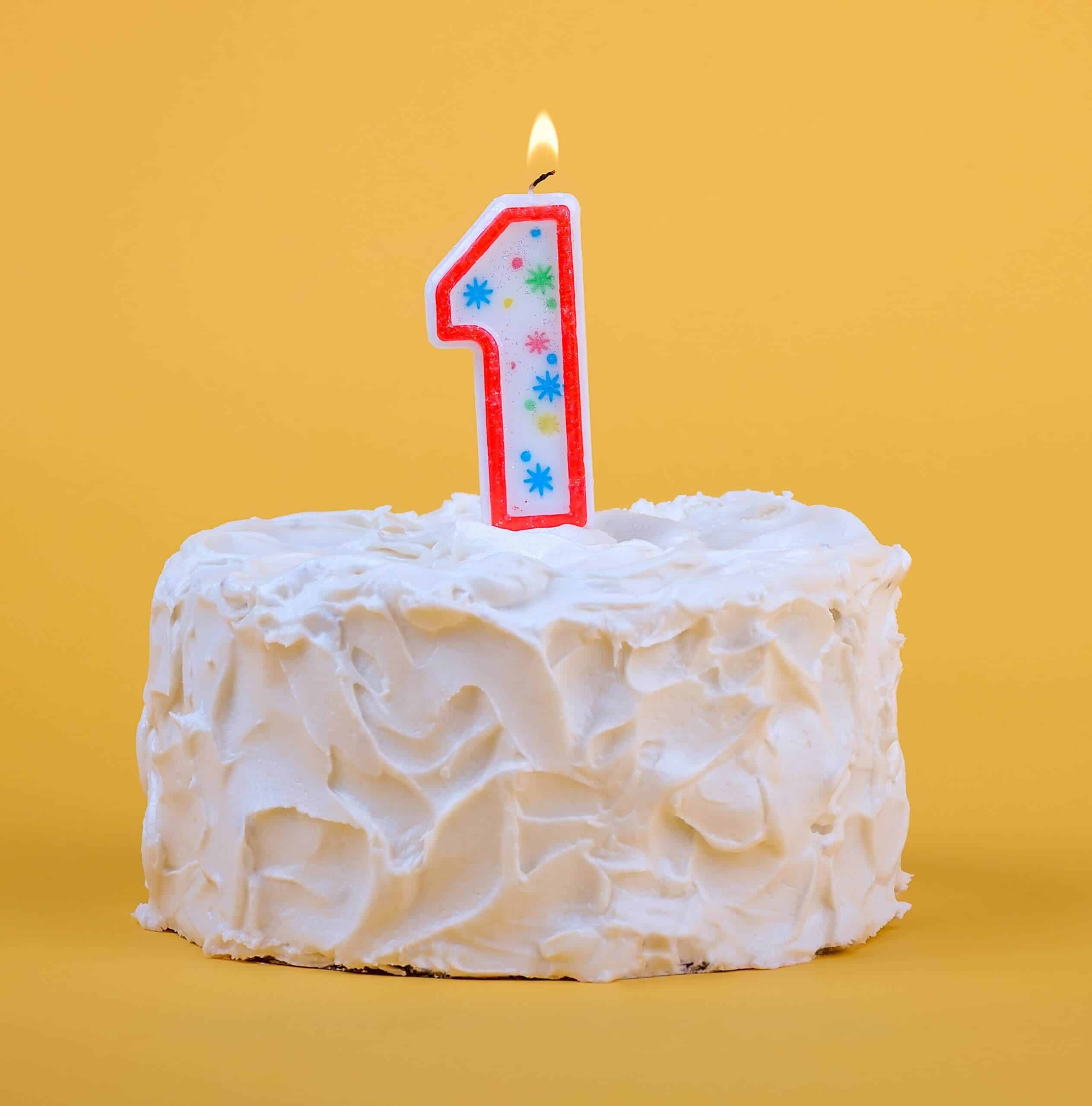 White frosted cake with a lit number 1 candle on a yellow background