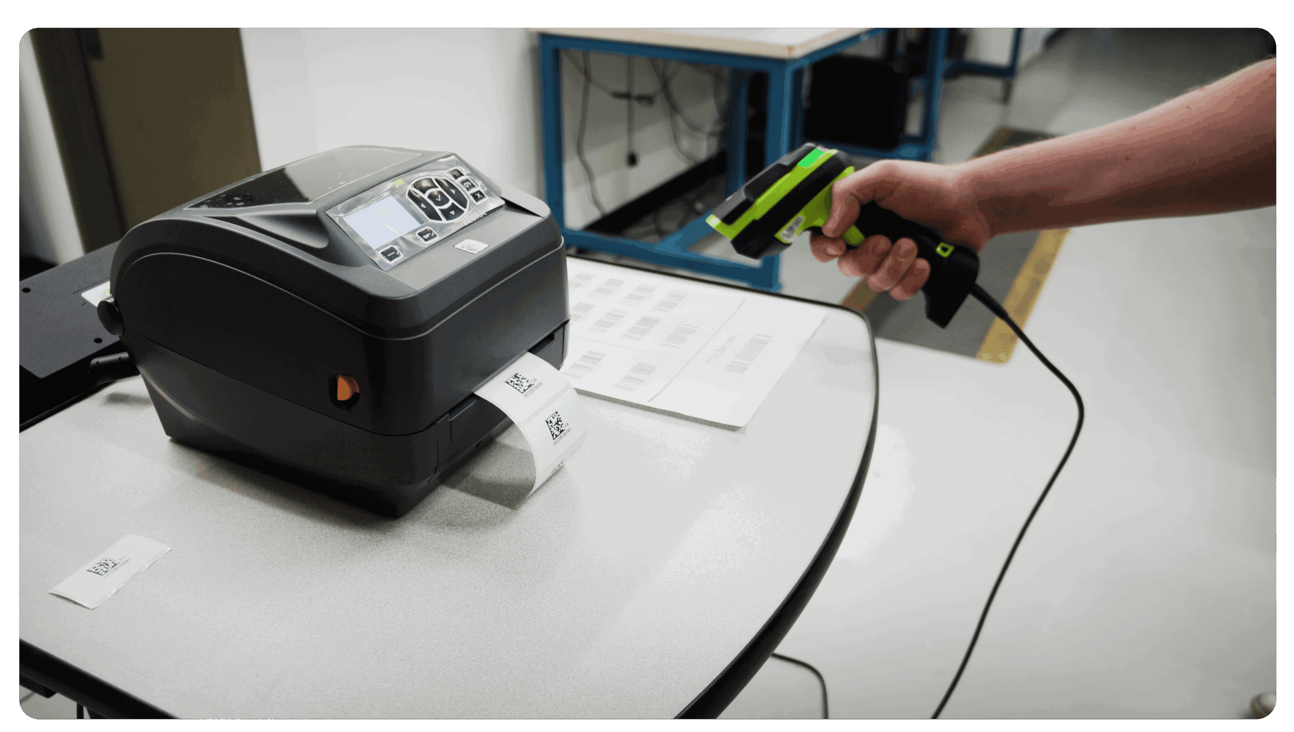 Hand scanning a barcode sheet with a handheld scanner next to a Zebra desktop label printer