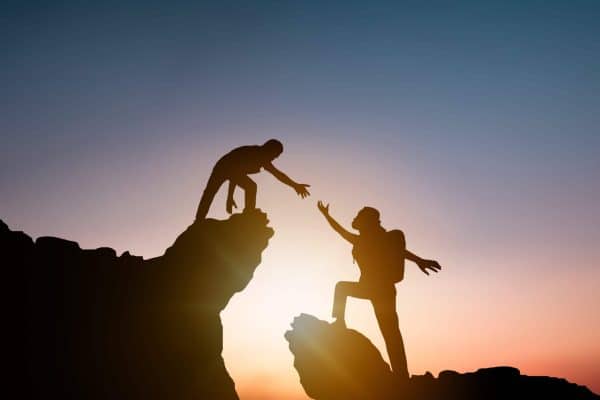 Silhouette of a hiker reaching down to help another climber up a cliff at sunset