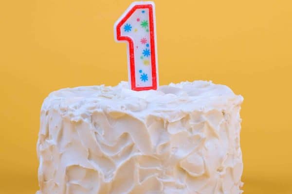 White frosted cake with a lit number 1 candle on a yellow background