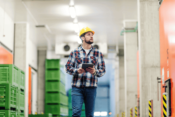 Surveyor in a hard hat carrying a tablet through a warehouse aisle of green crates
