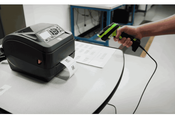 Hand scanning a barcode sheet with a handheld scanner next to a Zebra desktop label printer